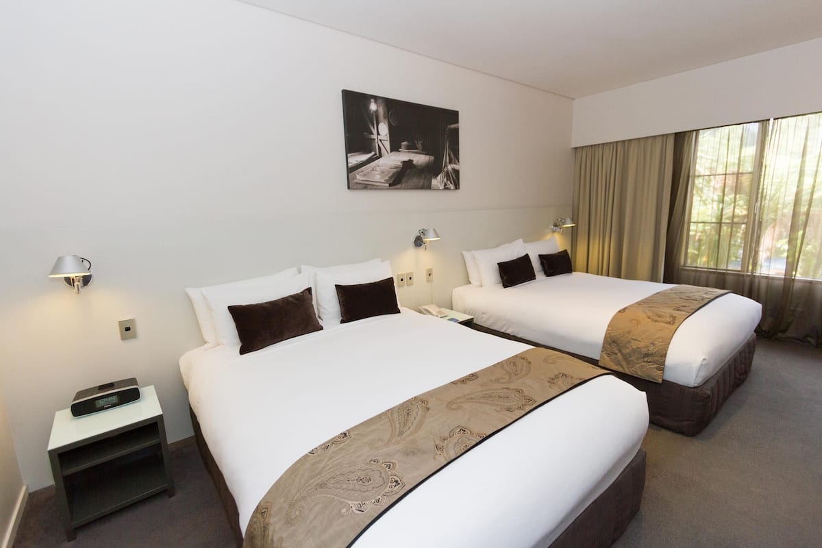 Una habitación de hotel con dos camas, una mesita de noche con un reloj, un cuadro en la pared y una ventana con cortinas.
