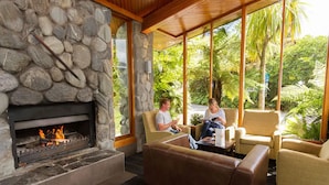 A stone fireplace with a fire, a wooden ceiling, and a view of lush greenery outside. - Scenic Hotel Franz Josef Glacier (Franz Josef Glacier)