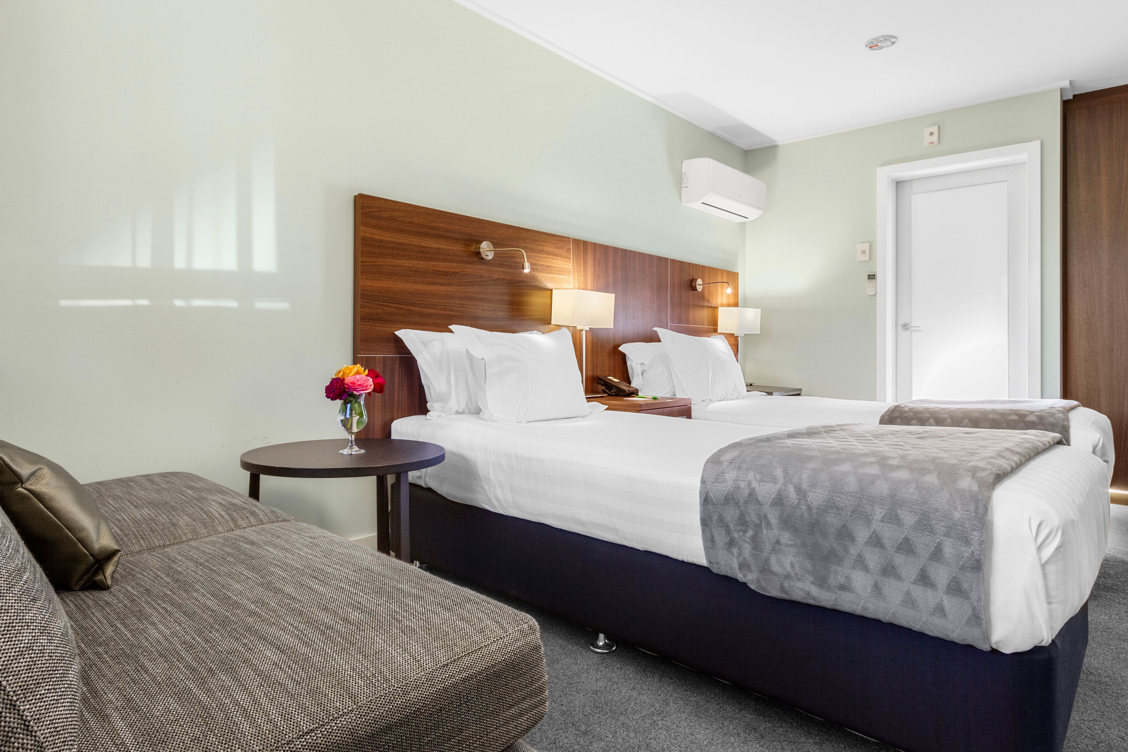 A hotel room with a large bed, two bedside tables, a small round table with a vase of flowers, a couch, and a door.