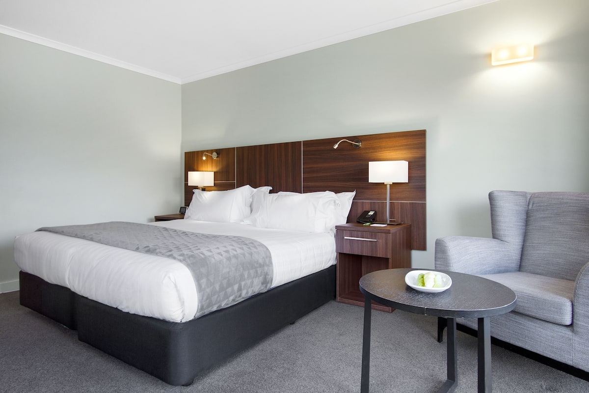 A hotel room with a large bed, a bedside table with a lamp, a chair, and a small table with a bowl of fruit.