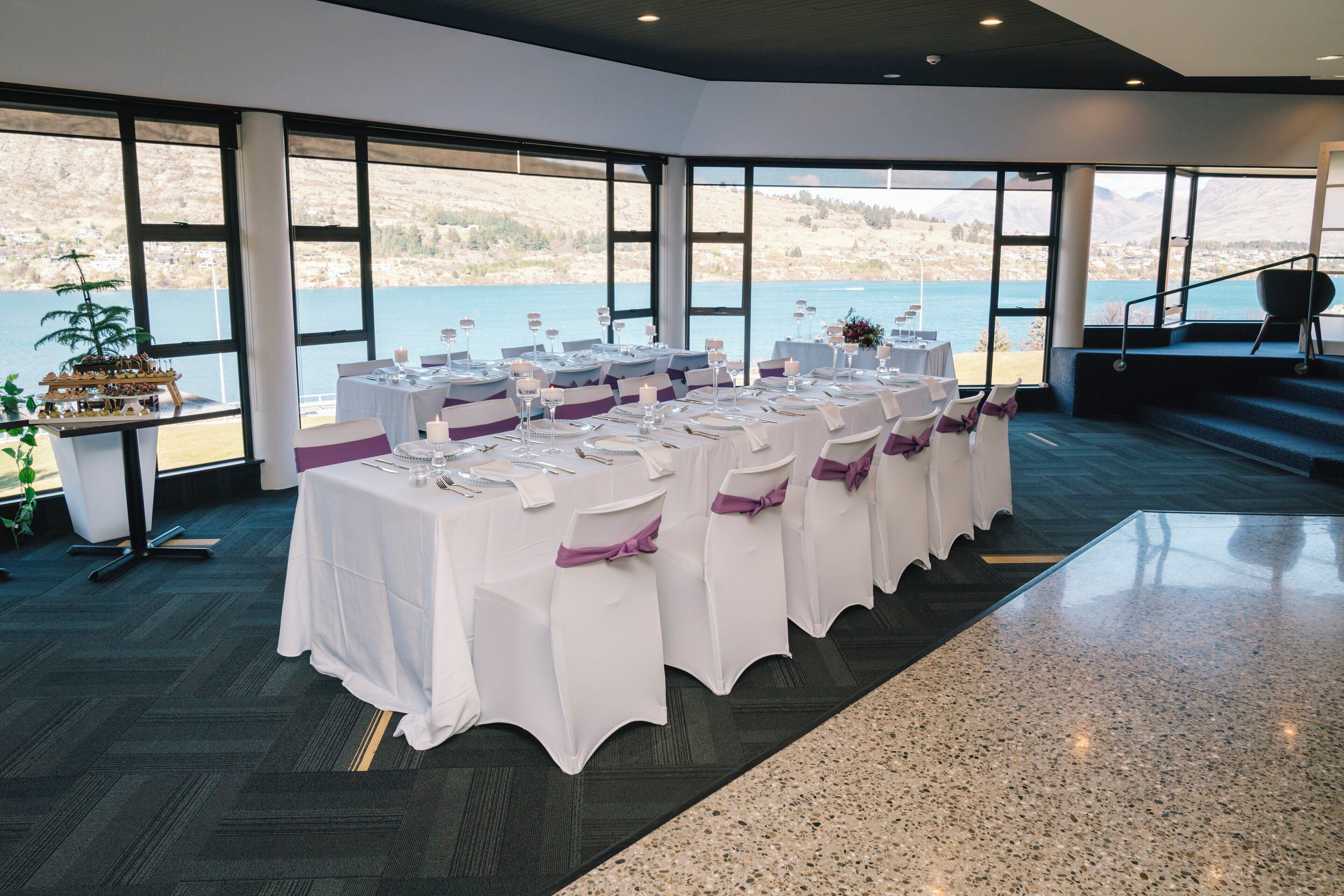 Un salón de banquetes con mesas dispuestas para un evento formal, grandes ventanales con vistas a un lago y montañas, y una escalera que conduce a un espacio exterior.