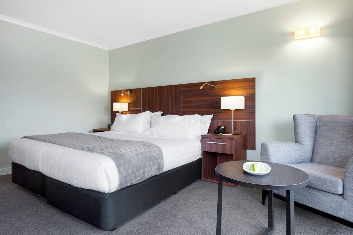 A hotel room with a large bed, a bedside table with a lamp, a small round table with a bowl, and a grey armchair.
