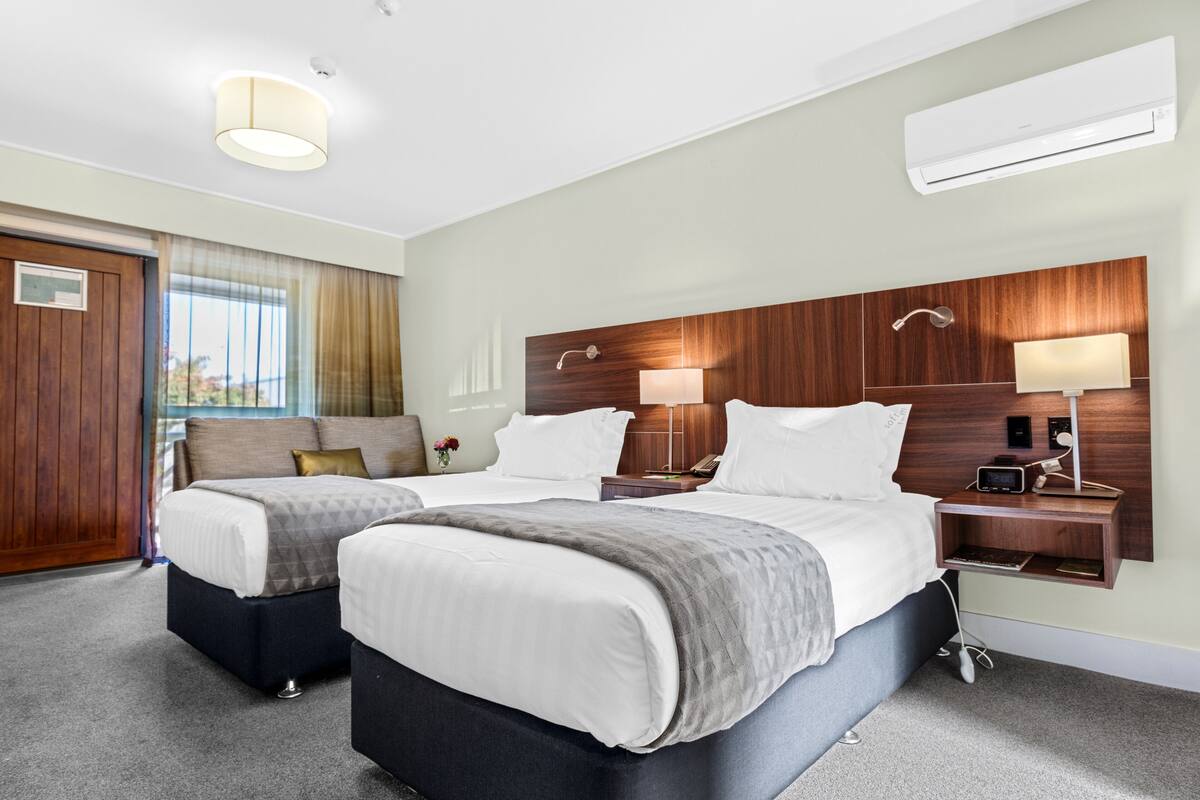 A hotel room with two beds, a wooden headboard, bedside lamps, an air conditioner, and a view of the outdoors.