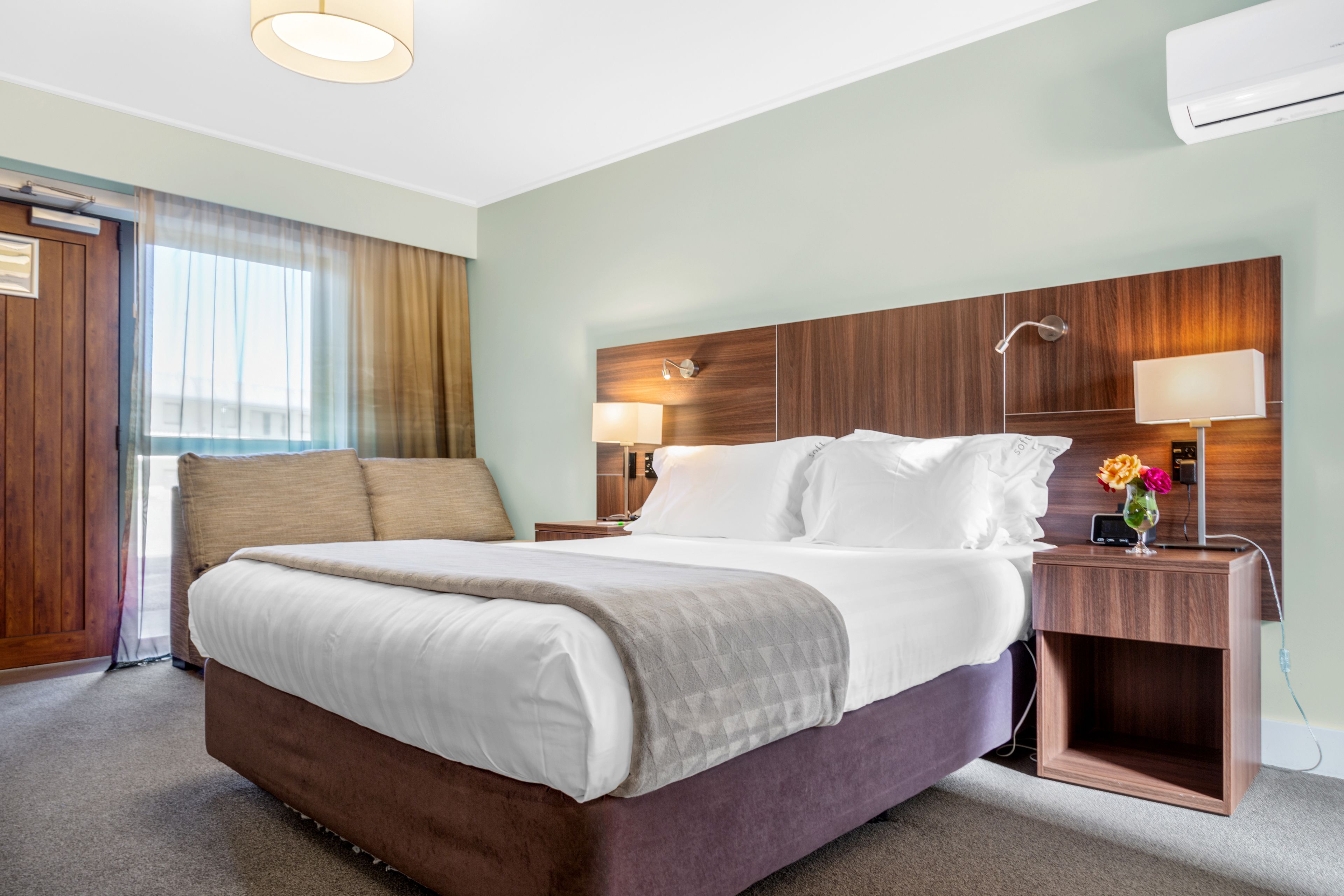 Quarto de hotel com uma cama grande, cabeceira de madeira, duas mesas de cabeceira com abajures e uma janela com cortinas.