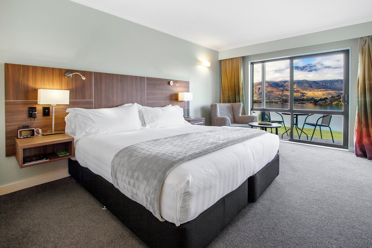 A hotel room with a large bed, a desk with a lamp, a chair, and a view of a lake and mountains.