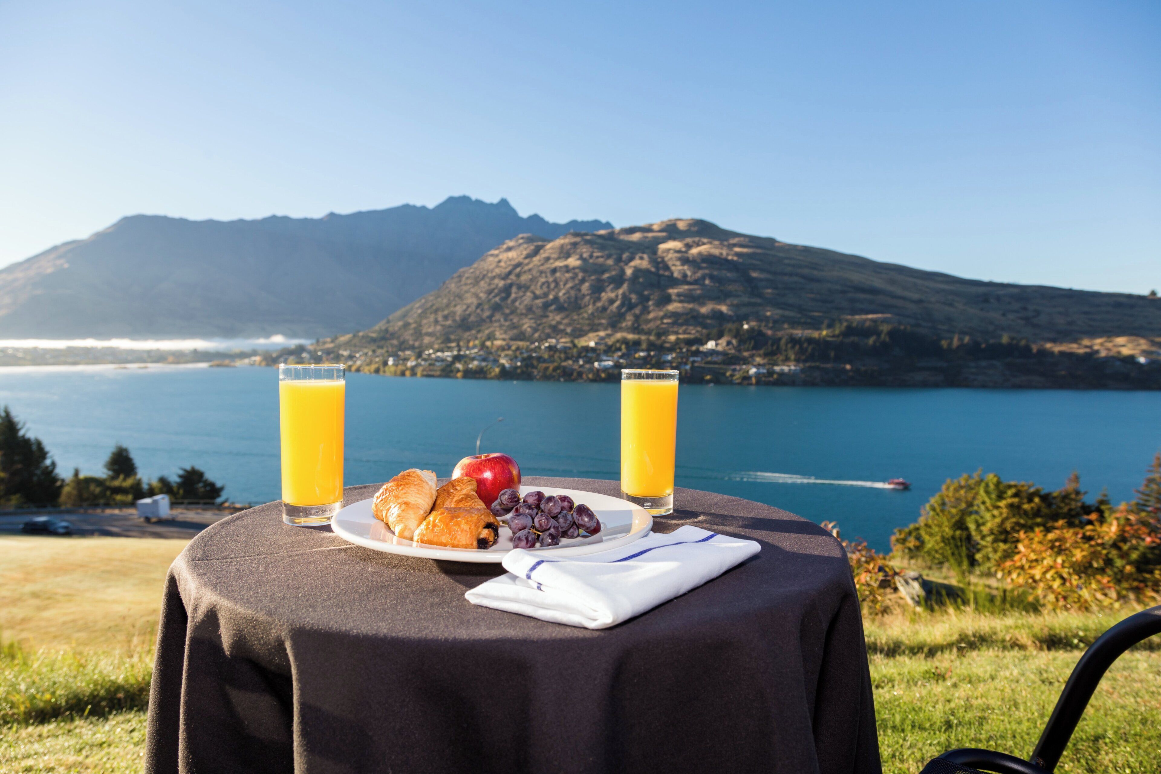 Una mesa con alimentos para el desayuno y dos vasos de jugo de naranja, con vistas a un lago y montañas.