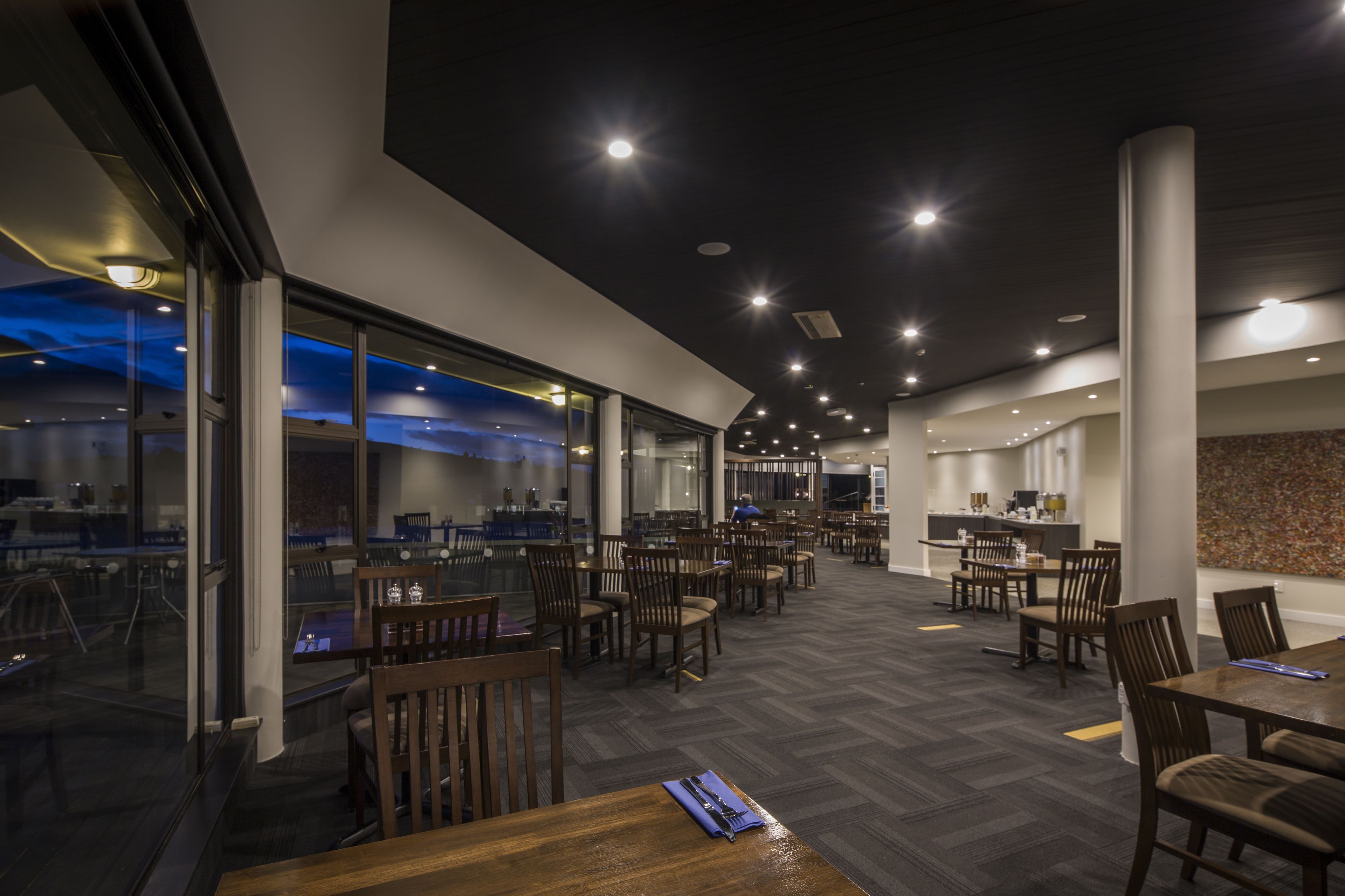 A modern dining area with wooden tables and chairs, large windows, and a high ceiling with recessed lighting.