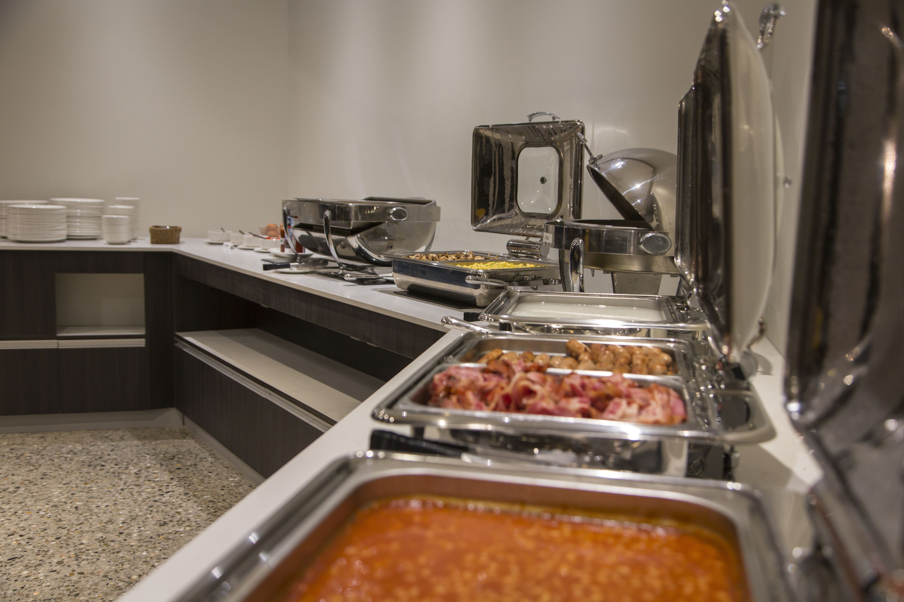 Een buffetopstelling met diverse soorten eten in metalen bakken, een koffieapparaat en een stapel witte borden op een toonbank.