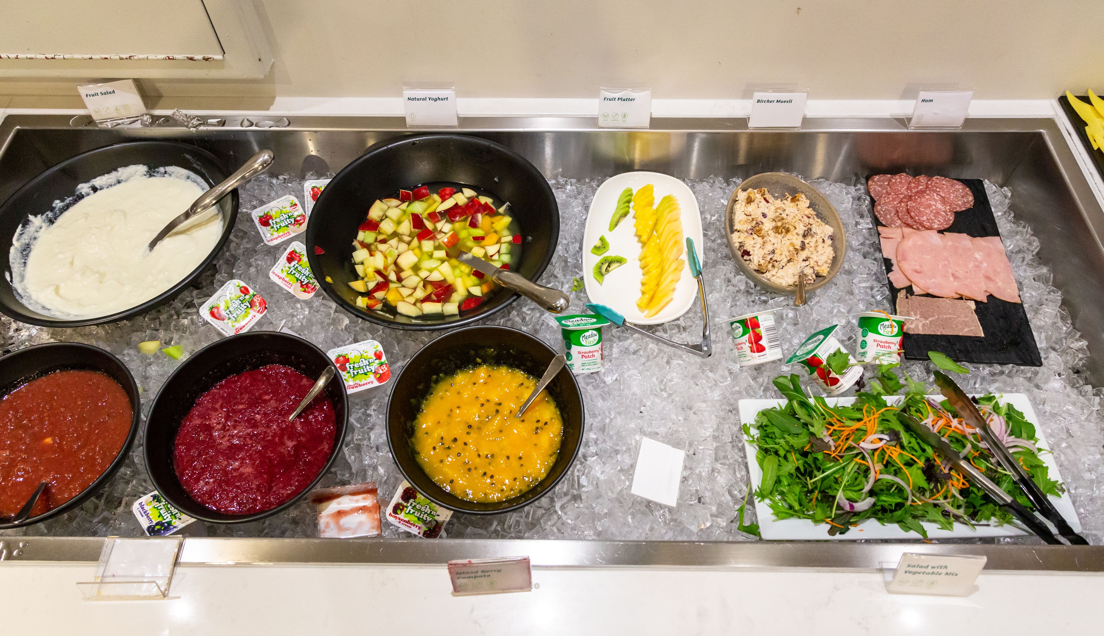 A buffet with various food options including fruit salad, natural yogurt, fruit platter, bircher muesli, ham, and a salad with vegetable mix.