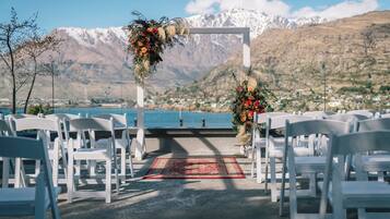 A wedding ceremony setup with white chairs arranged in rows, a red carpet, and floral decorations against a backdrop of mountains and a lake.