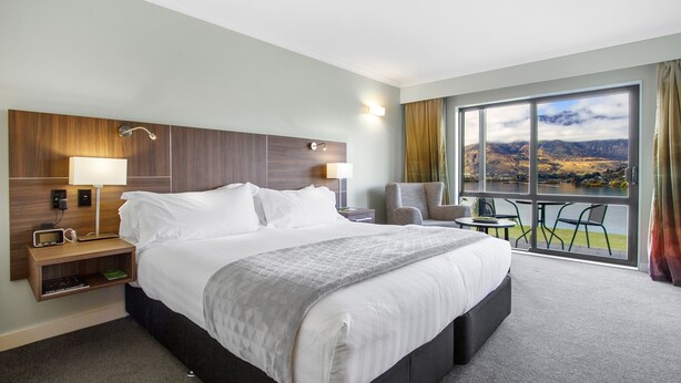 A hotel room with a large bed, a desk with a lamp, a chair, and a view of a lake through a large window. - Holiday Inn Queenstown Frankton Road by IHG (Queenstown)
