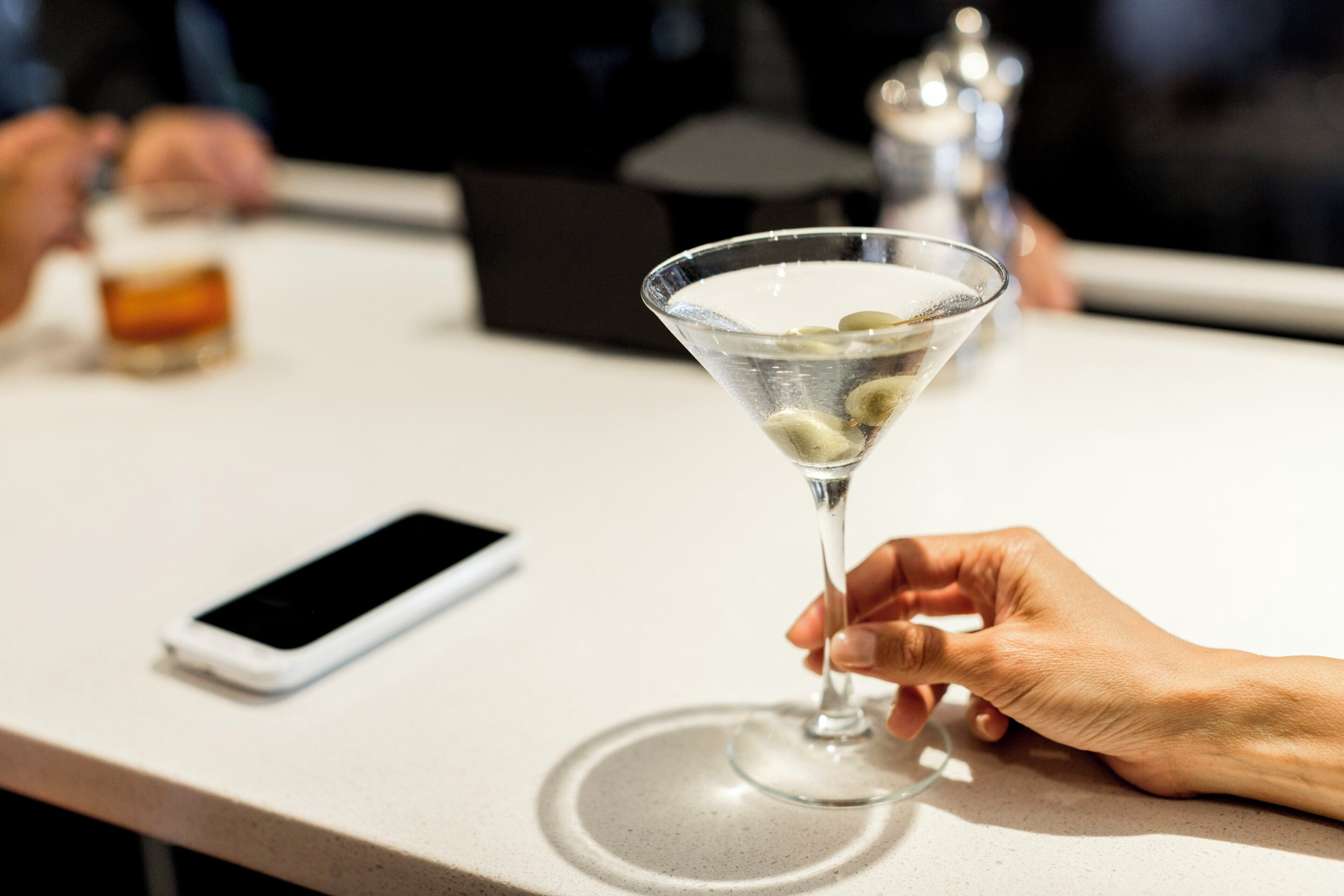 A martini glass with ice cubes on a bar counter, a smartphone, and a blurred background suggesting a social setting.