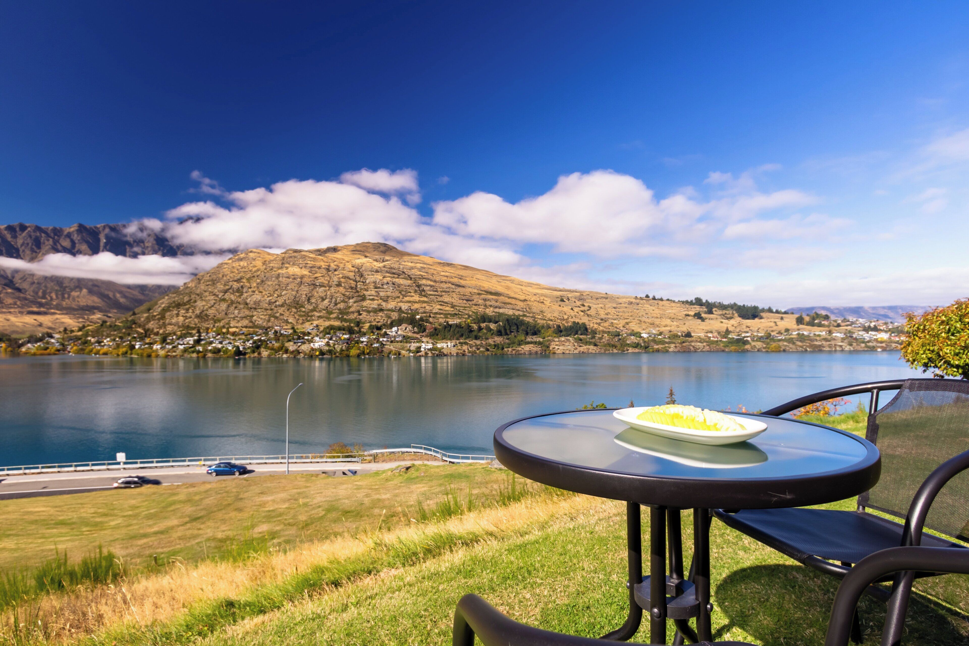 A lakeside view with a mountain in the background, a dining table with a plate of food, and a chair.