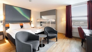 Habitación de hotel con cama, dos sillones, escritorio, ventana grande con cortinas, proyector de techo y vista a la ciudad.