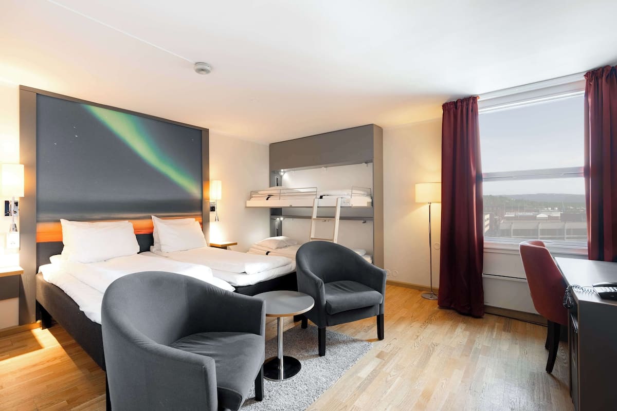 A hotel room with a bed, two armchairs, a desk, a large window with curtains, a ceiling projector, and a view of the city.