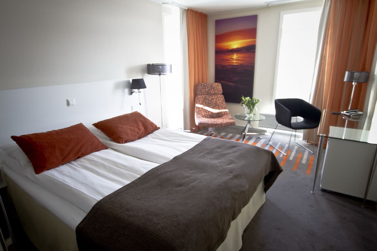 A hotel room with a bed, a desk, a chair, a TV, and a painting of a sunset.