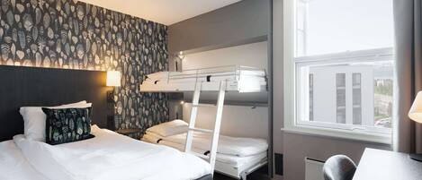 A hotel room with a bunk bed, a desk, a chair, a window with a view, and a patterned wall.