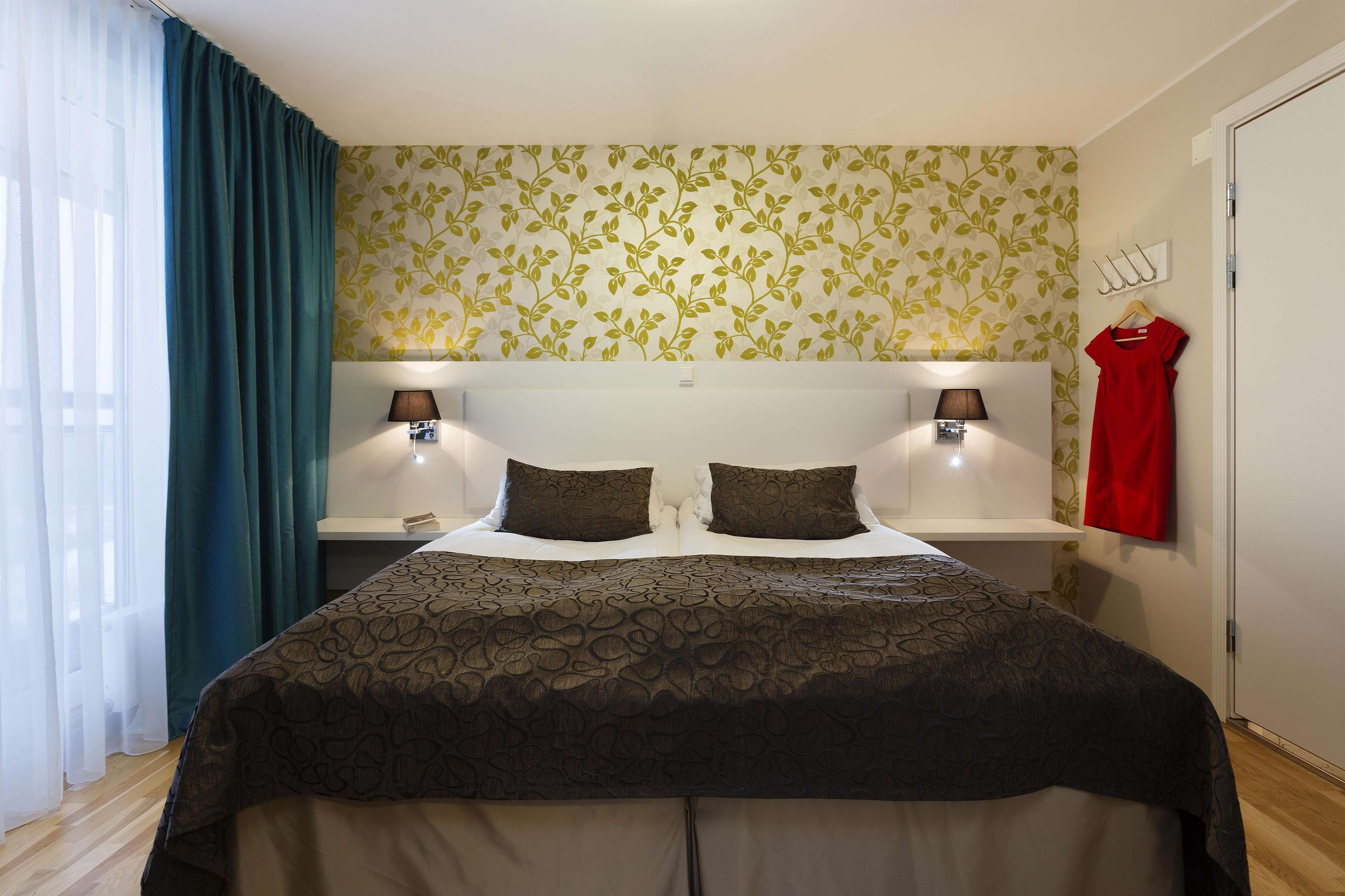 Ein ordentlich bezogenes Bett mit einem gemusterten Überwurf, zwei Nachttische mit Lampen, ein rotes Kleid an einem Kleiderbügel und eine blumige Tapete.