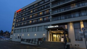 Un hotel moderno con un letrero que dice "Scandic Alta". Cuenta con varios pisos con balcones, una entrada bien iluminada y un estacionamiento visible.