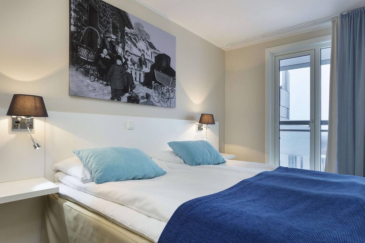 A hotel room with a bed, two bedside lamps, a large black-and-white photo on the wall, and a window with curtains.