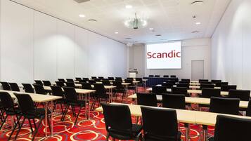 A conference room with rows of tables and chairs, a projector screen with the word "Scandic" on it, and a carpeted floor.