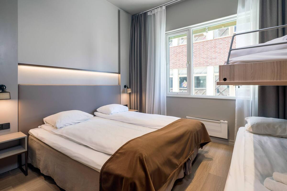 A hotel room with a bunk bed, a single bed, a bedside table with a lamp, and a window with curtains.