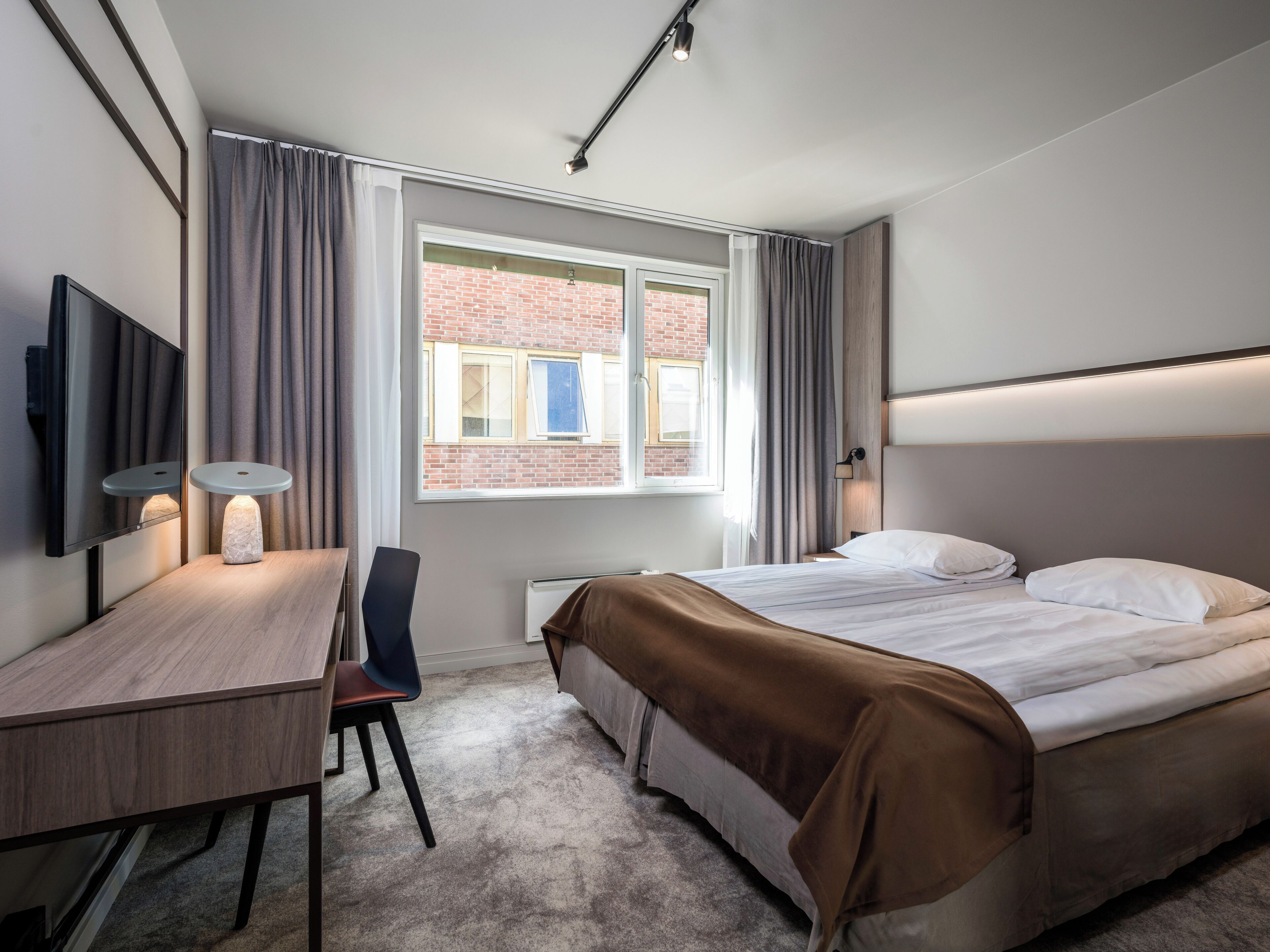 Moderni hotellihuone, jossa on sänky, työpöytä, tuoli, televisio ja ikkuna, jossa on verhot.