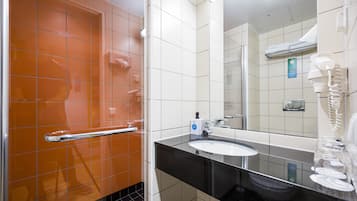 Un baño con un lavamanos negro, azulejos blancos y naranja, cabina de ducha con mampara de vidrio y un secador de manos.