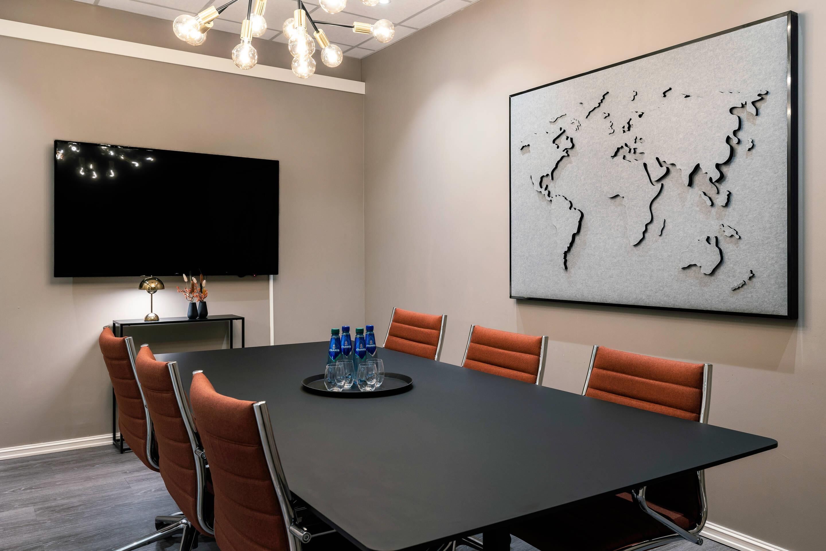 A conference room with a large table, six chairs, a flat-screen TV, a world map artwork, and a chandelier.