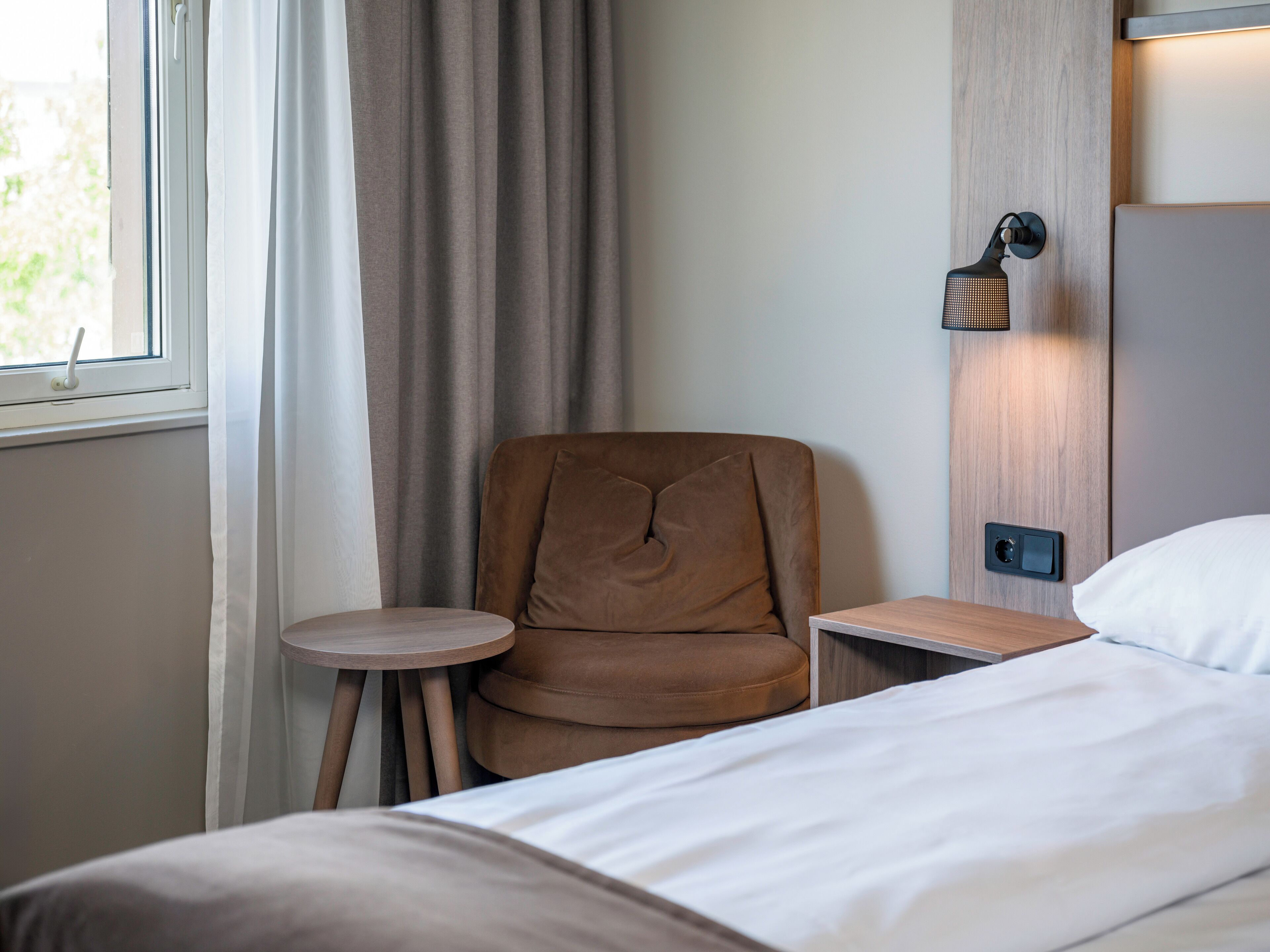 A hotel room with a bed, a small round table, a brown chair, and a wall-mounted lamp.