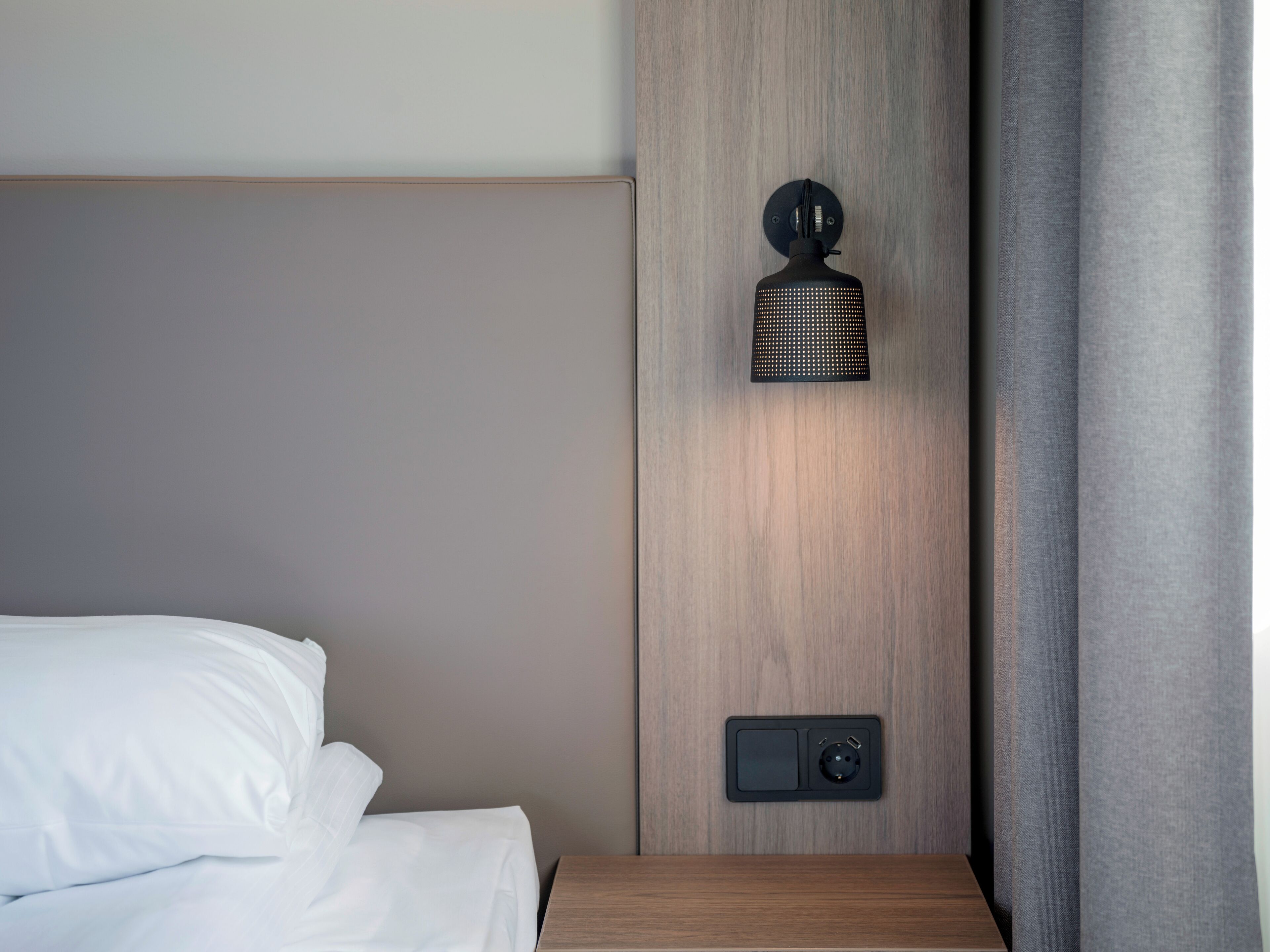 Una cama bien tendida con sábanas blancas, una mesita de noche de madera, una lámpara de pared y un enchufe.