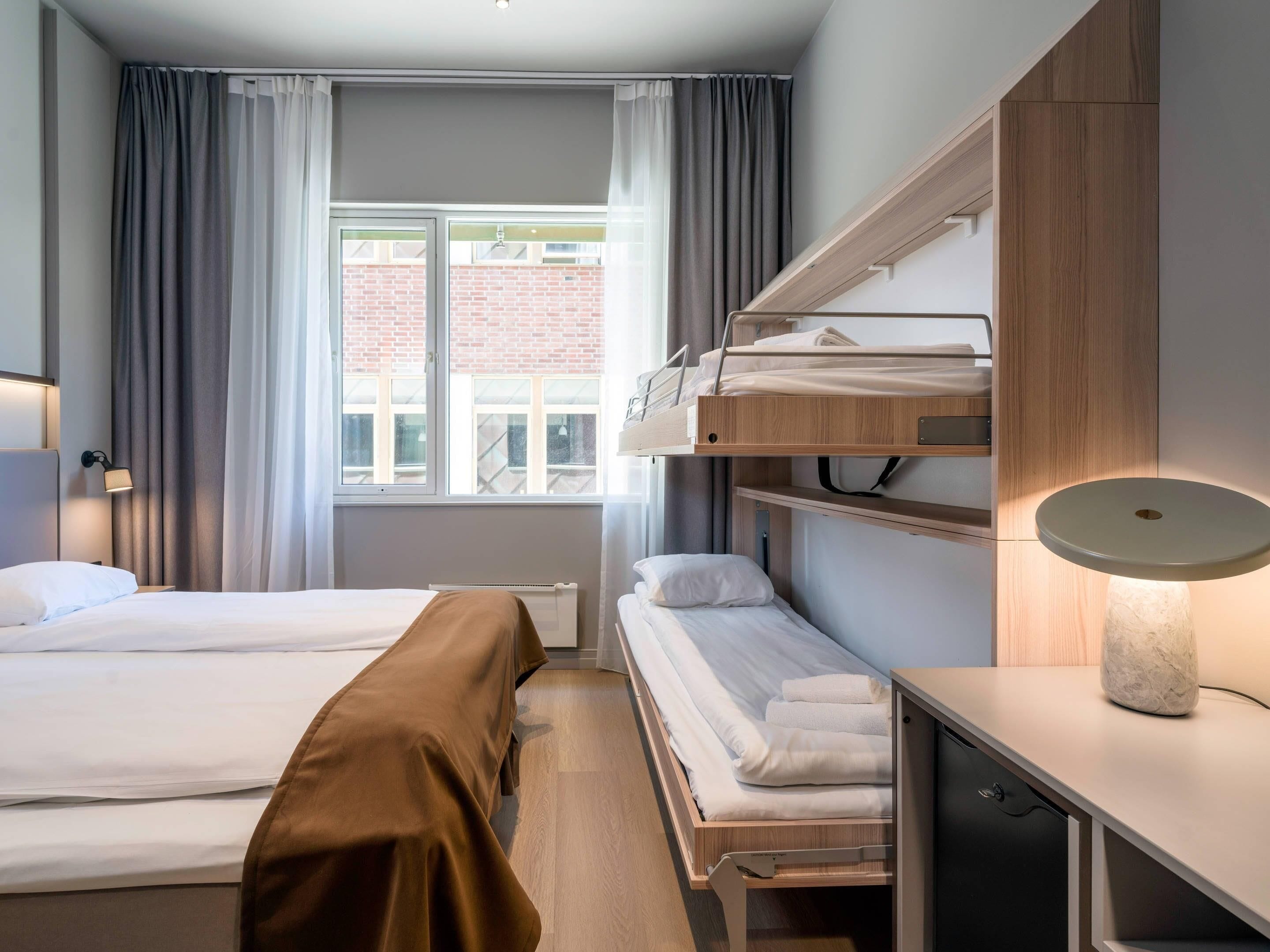 Ein Hotelzimmer mit einem Bett, einem Schreibtisch mit Lampe, einem Fenster mit Vorhängen und Blick auf ein Backsteingebäude.