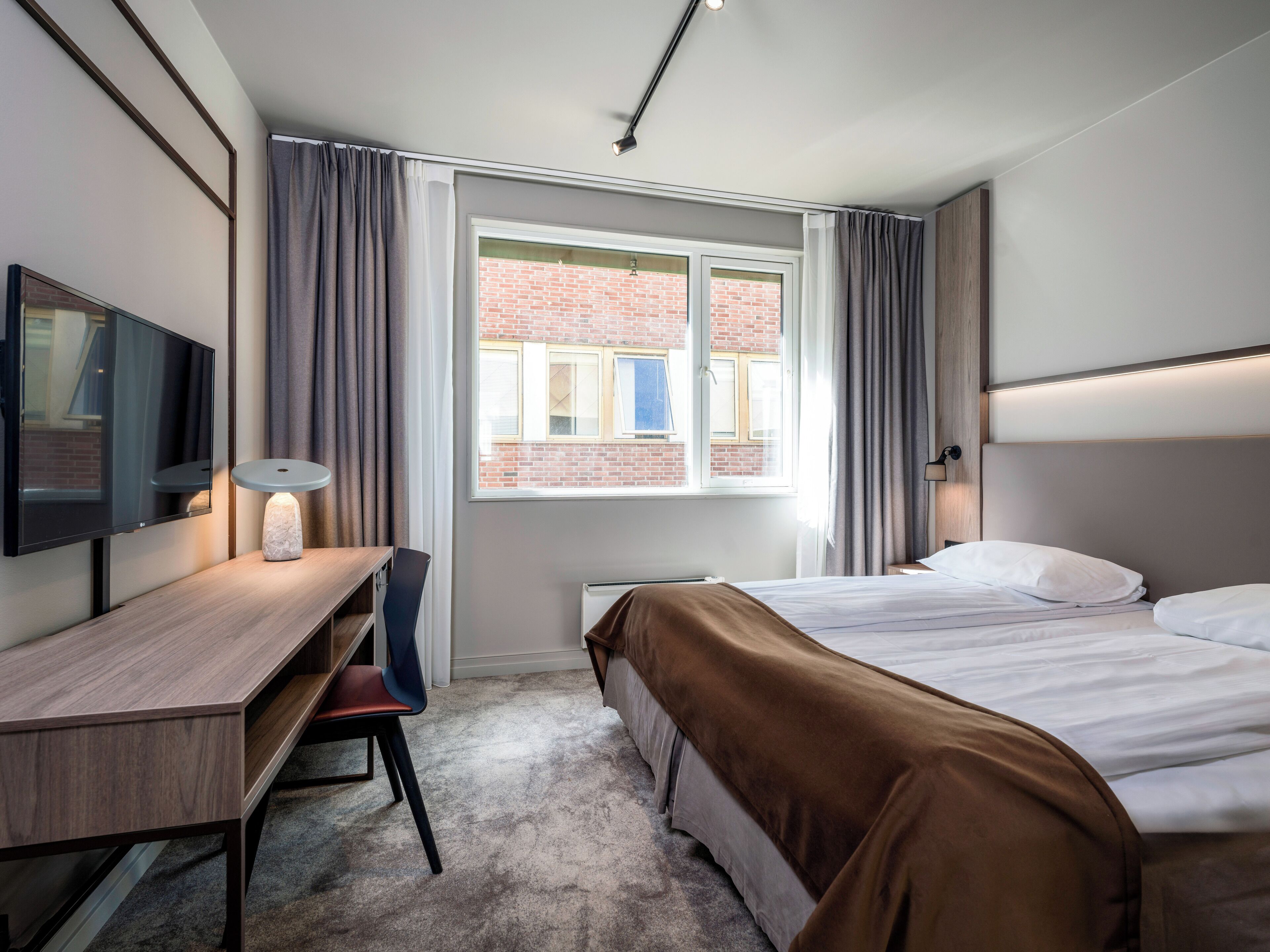 A hotel room with a bed, a desk with a chair, a TV, and a window with curtains.