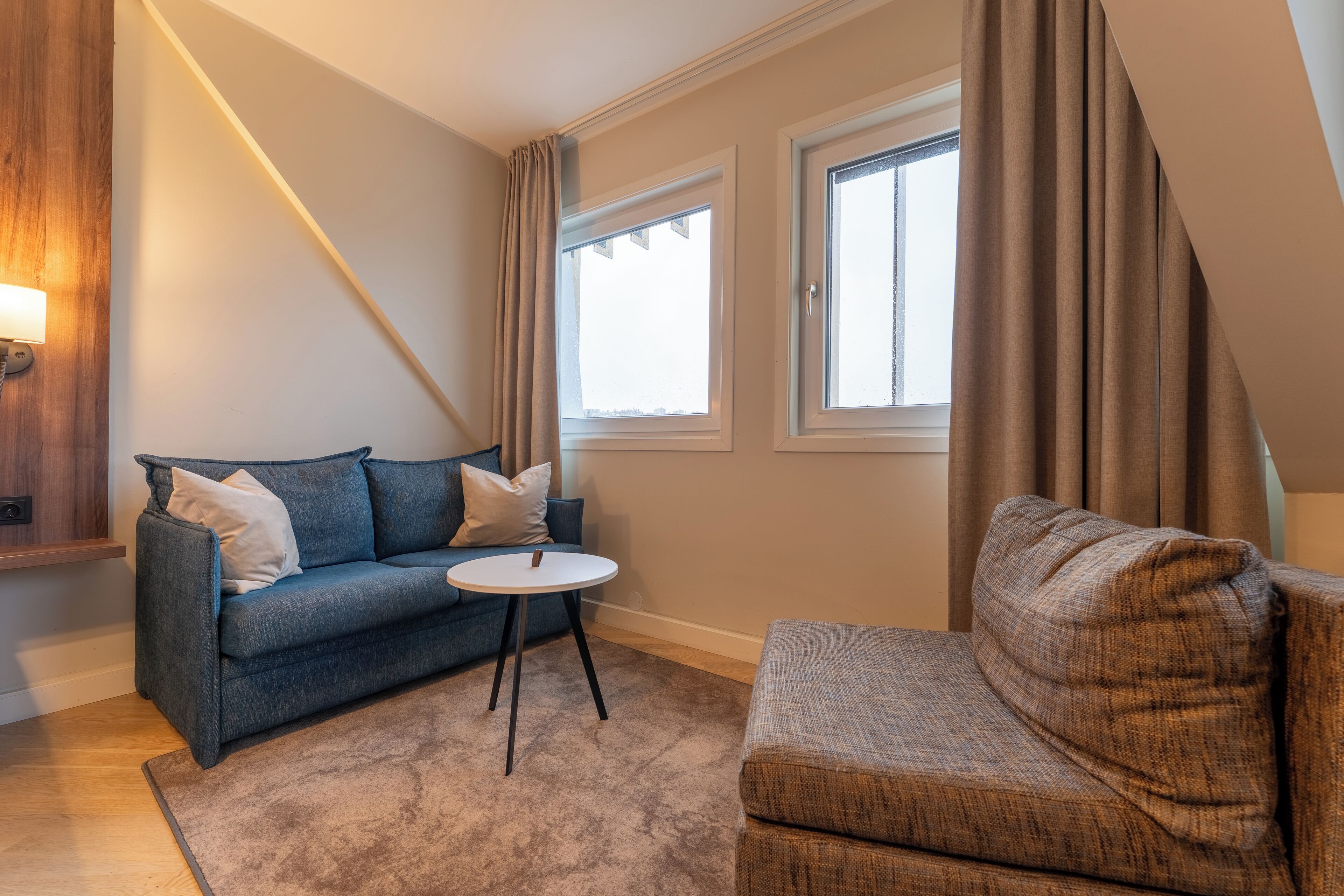 A cozy living room with a blue sofa, a brown armchair, a small round table, and two windows with curtains.