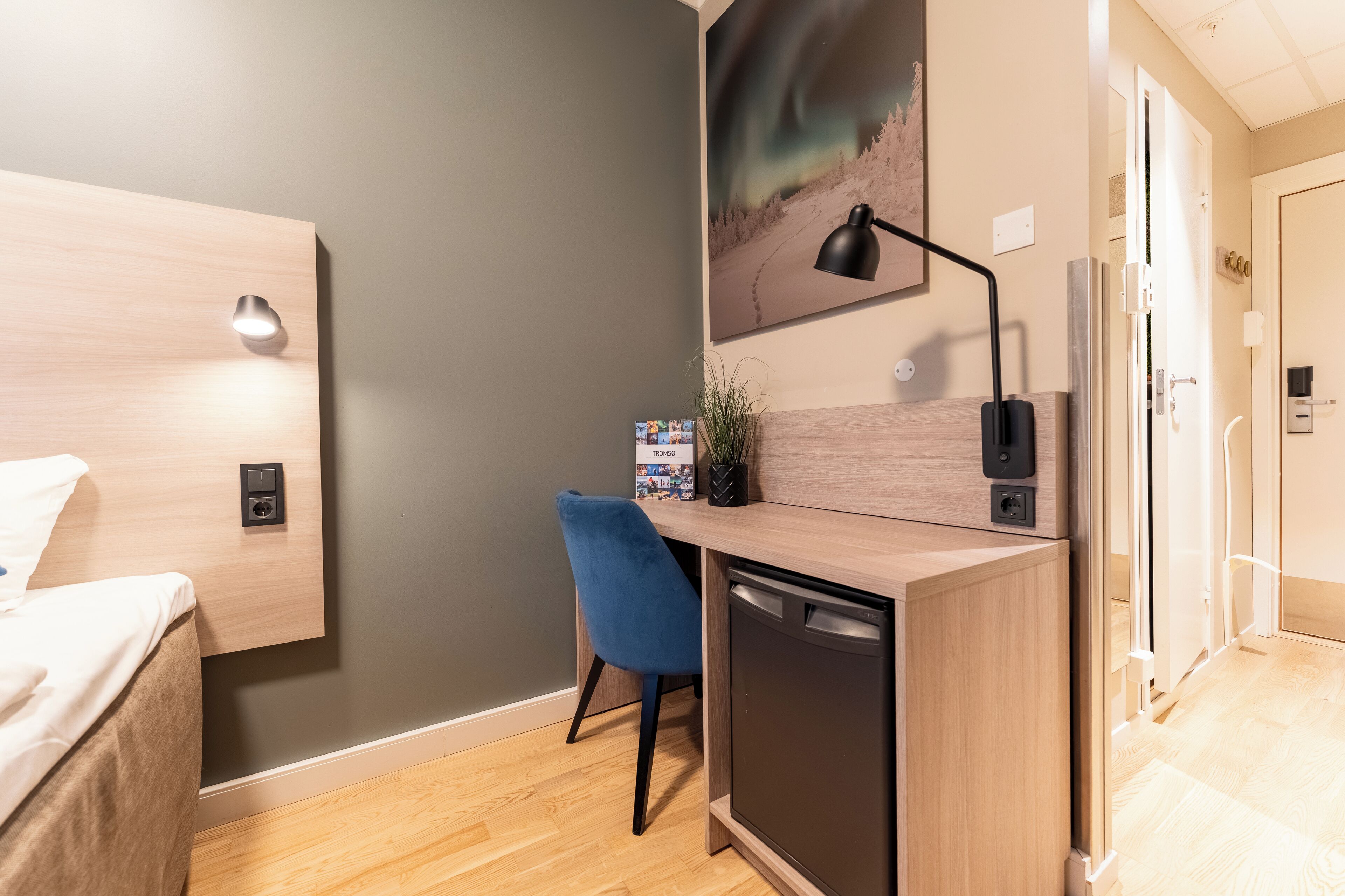 A modern hotel room with a wooden desk, a blue chair, a bed, a wall-mounted lamp, and a painting of the aurora borealis.