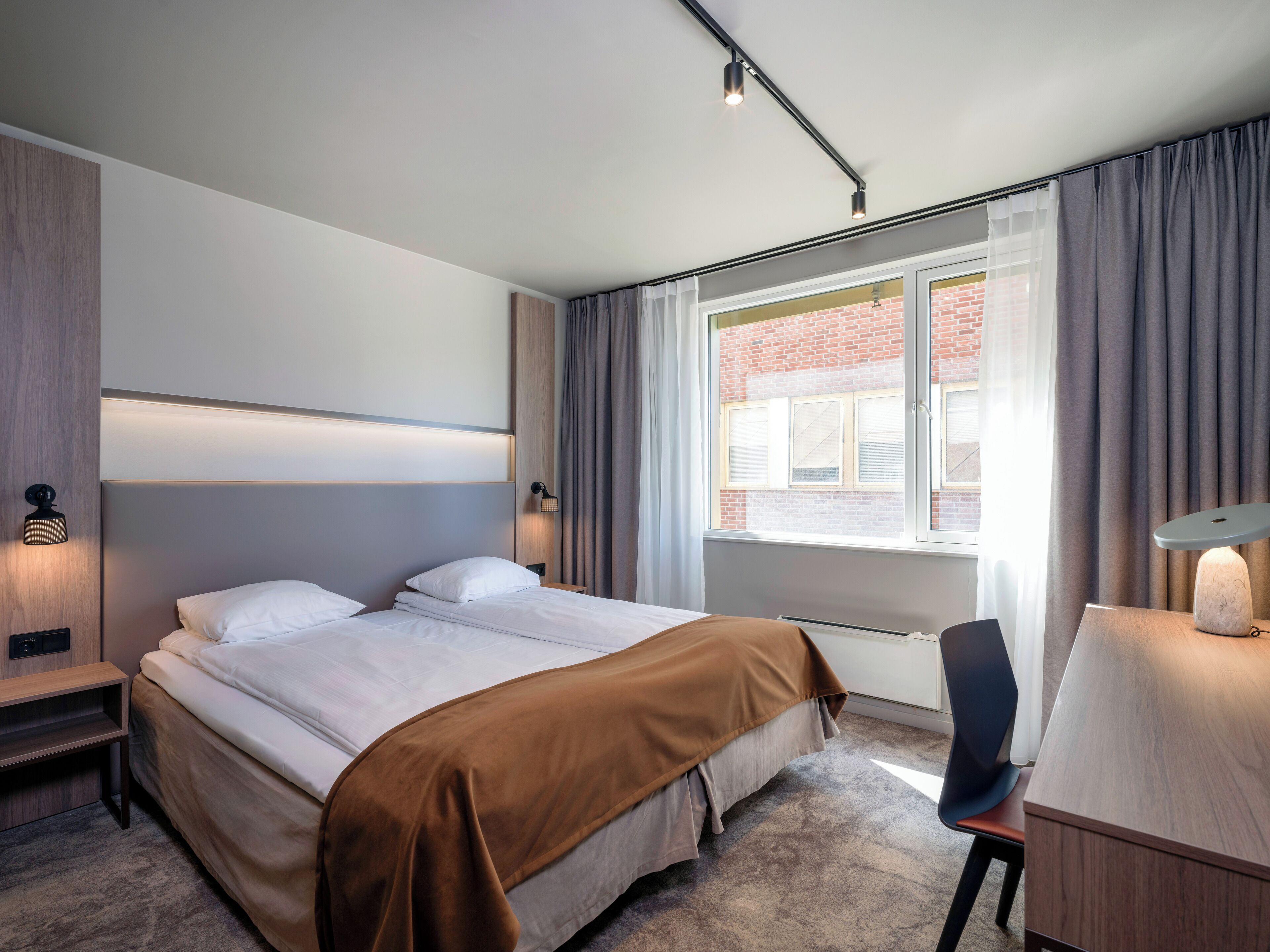A modern hotel room with a large bed, bedside lamps, a desk with a chair, and a window with curtains.