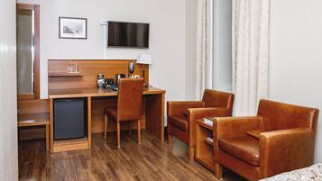 A hotel room with a wooden desk, two leather chairs, a small fridge, a TV, and a bed with a patterned bedspread.