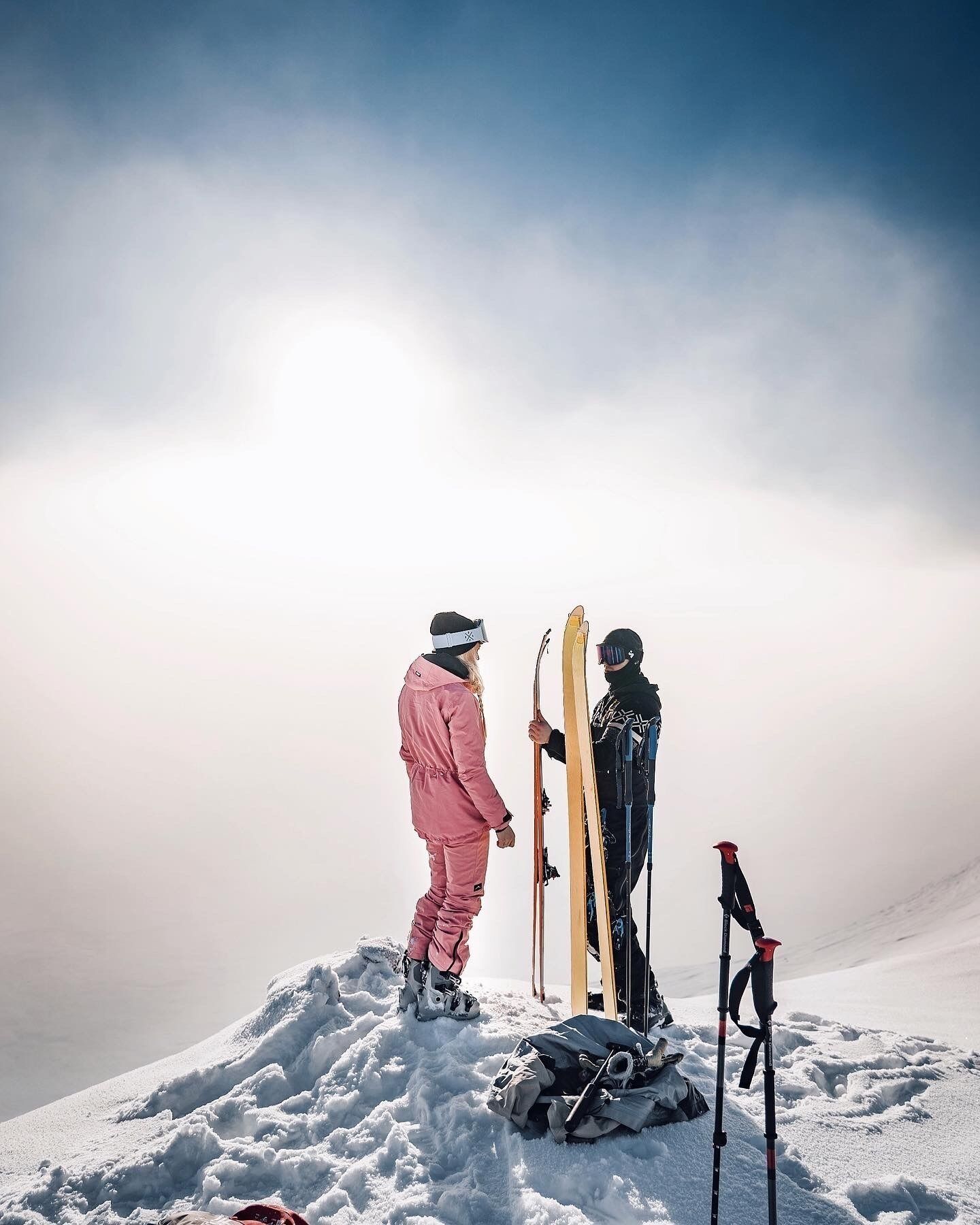 Dos personas en la cima nevada, con esquíes, una sosteniendo un par de esquíes amarillos y la otra un par de esquíes negros.