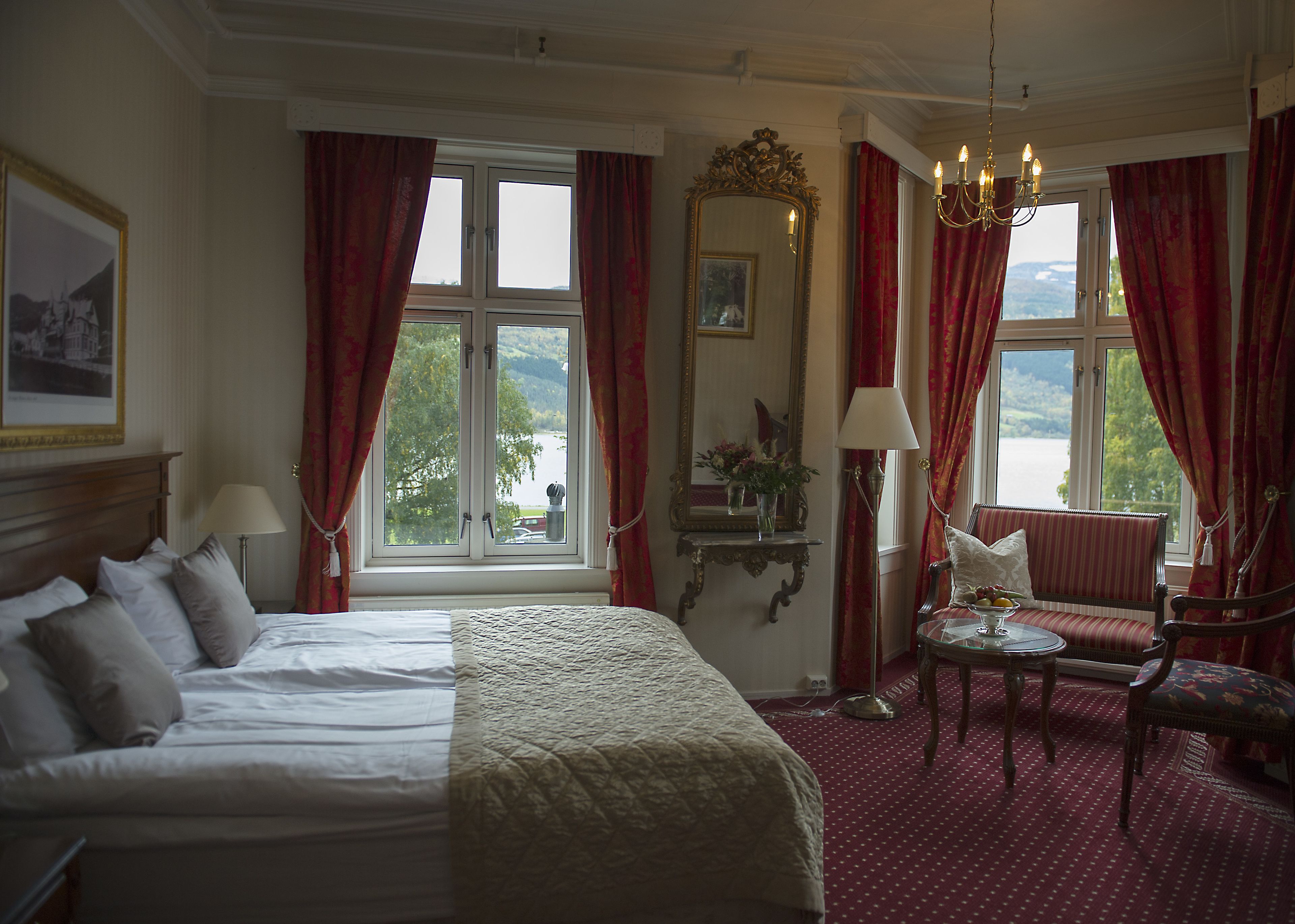 A hotel room with a bed, a chair, a table with a vase, a mirror, and a view of the outdoors.
