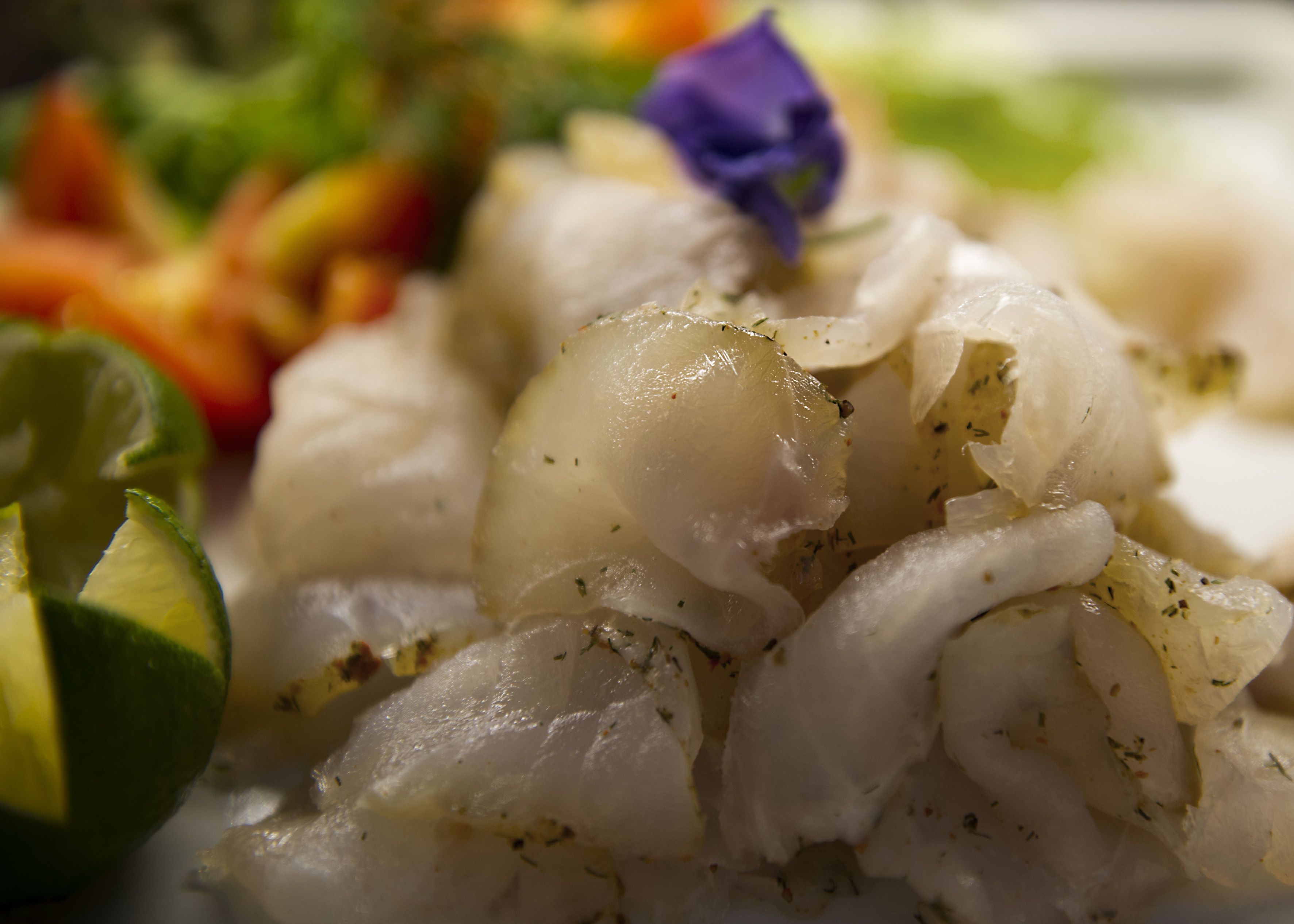Primer plano de un plato con pescado blanco, hierbas y una flor púrpura como adorno.