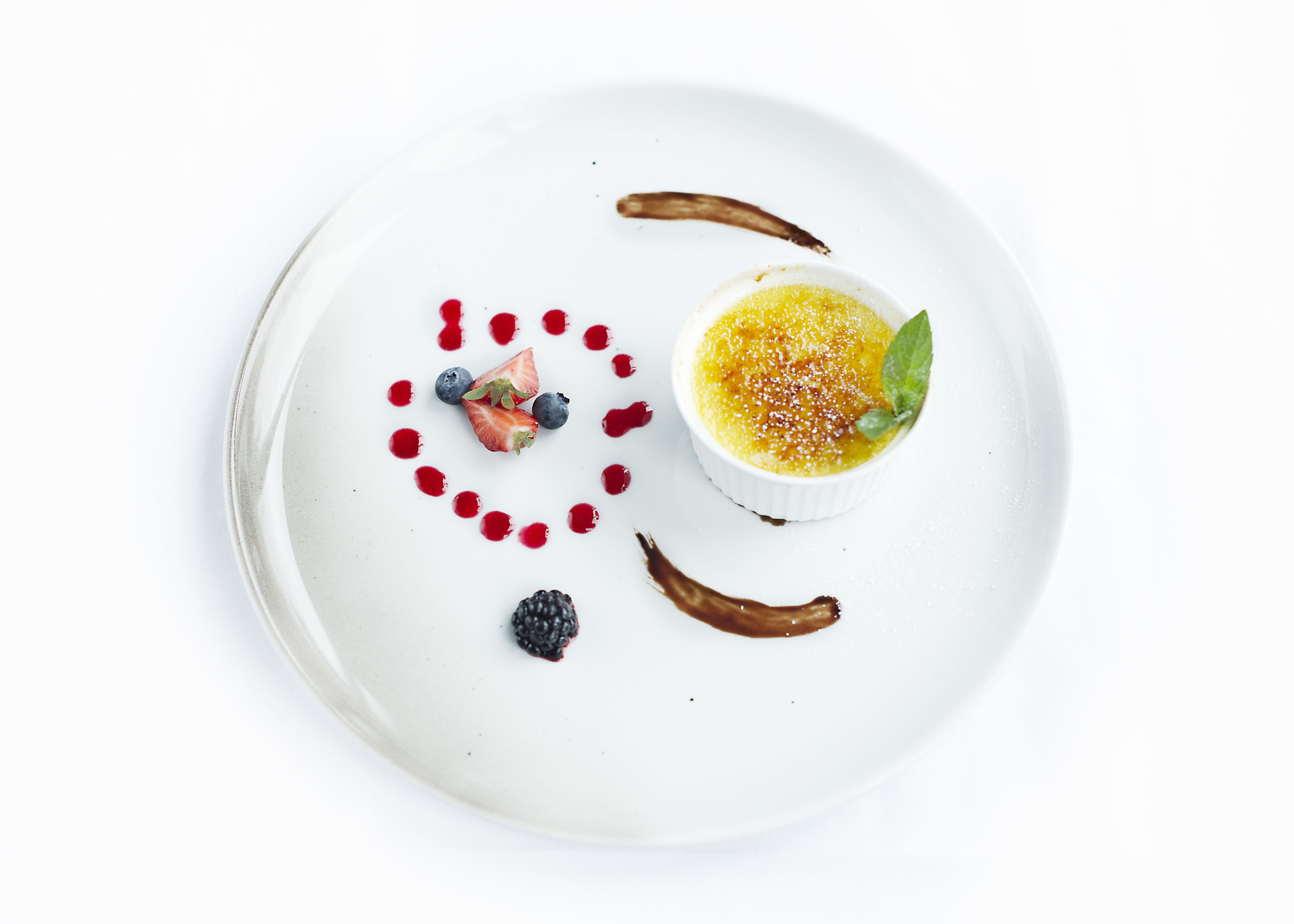 Un plato de postre con crema pastelera, frutos rojos y salsa de chocolate.