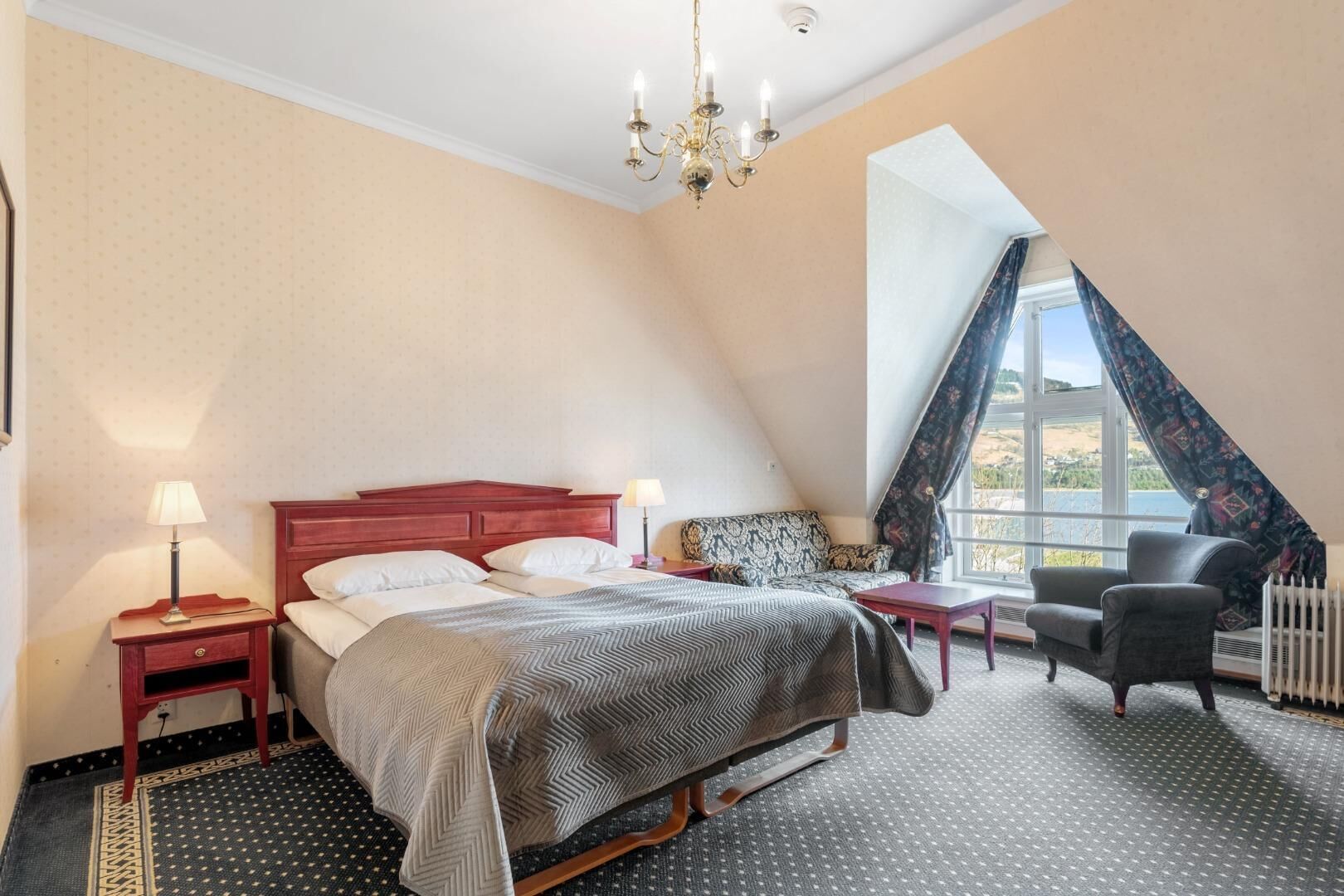 Ein Schlafzimmer mit Bett, Nachttischen, einem Sessel, einem Fenster mit Vorhängen und einer Pendelleuchte.
