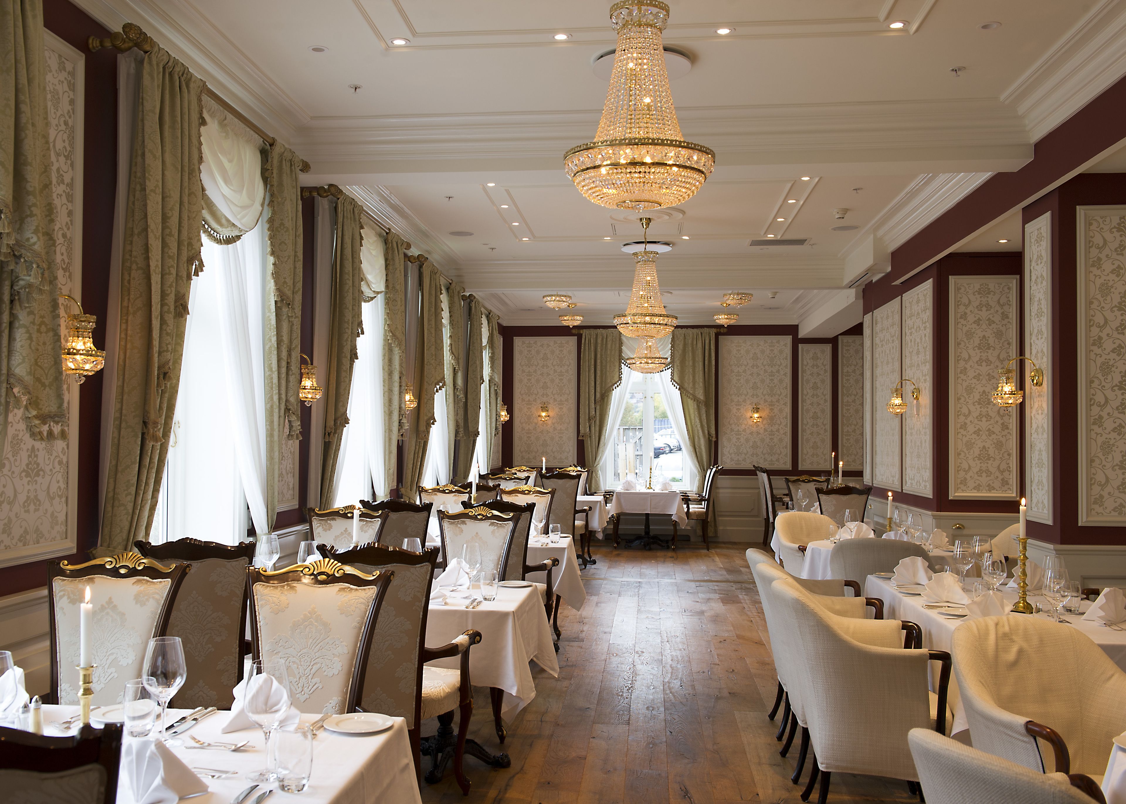 A formal dining room with multiple tables and chairs, ornate chandeliers, and elegant wall decorations.