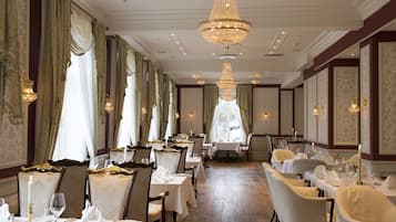 A formal dining room with multiple tables and chairs, ornate chandeliers, and elegant wall decorations.