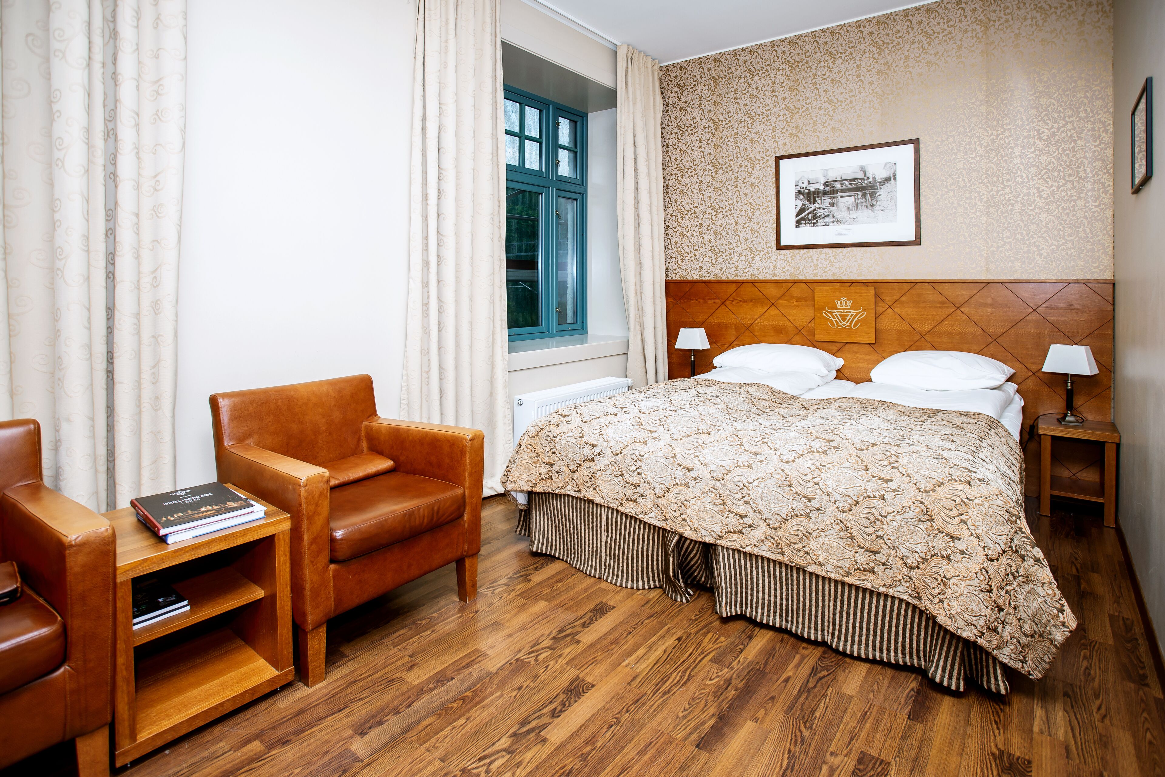 A hotel room with a wooden bed, a wooden chair, a small table, a window with curtains, a framed picture on the wall, and a radiator.