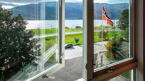A view from a window overlooking a lake with mountains in the background, a flag, and a road with vehicles. - Fleischer's Hotel (Voss)