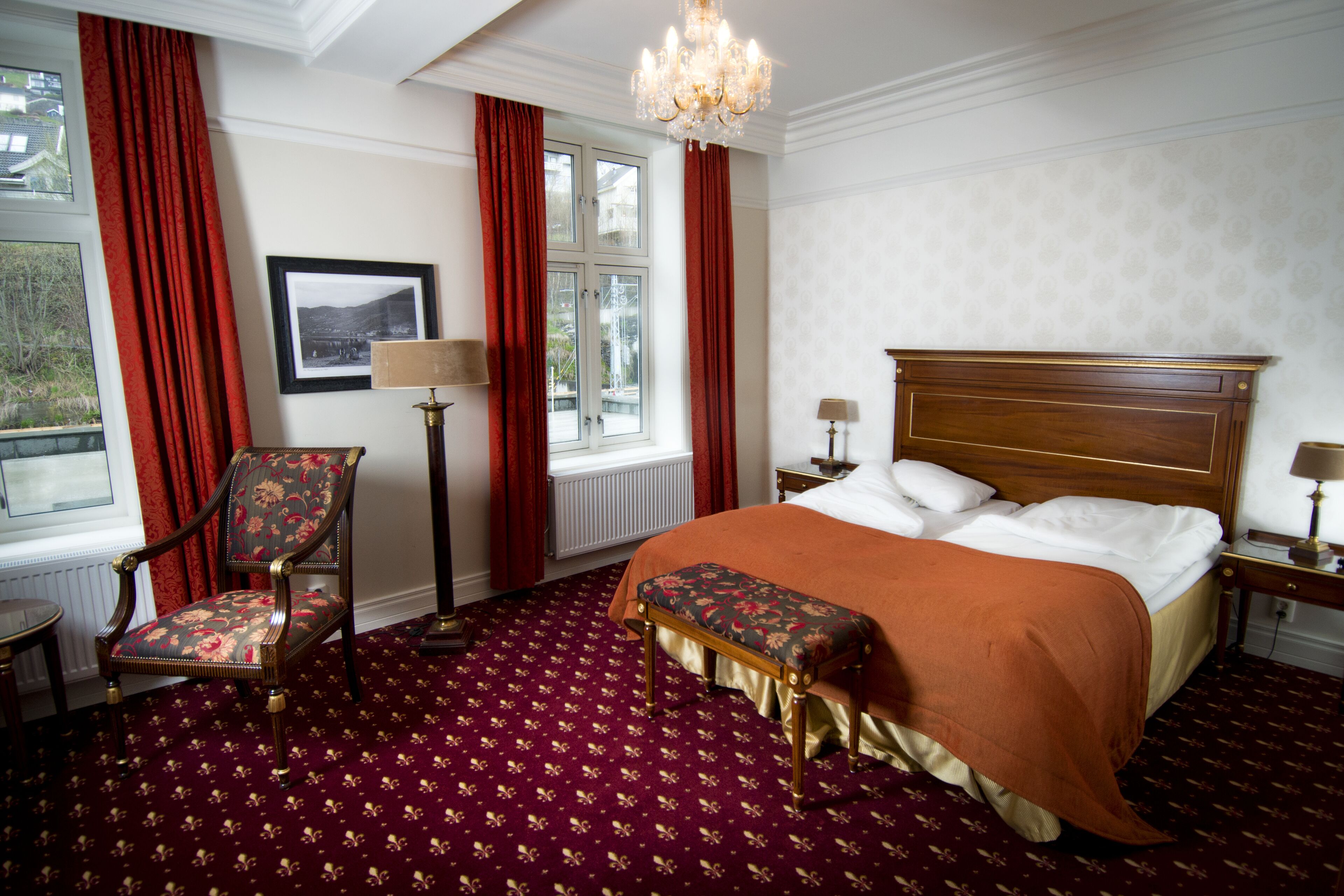 Habitación de hotel con una cama grande, cabecera de madera, alfombra roja, una silla, una lámpara de pie y una ventana con cortinas rojas.
