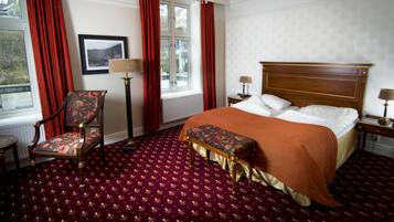 A hotel room with a large bed, a wooden headboard, a red carpet, a chair, a floor lamp, and a window with red curtains.