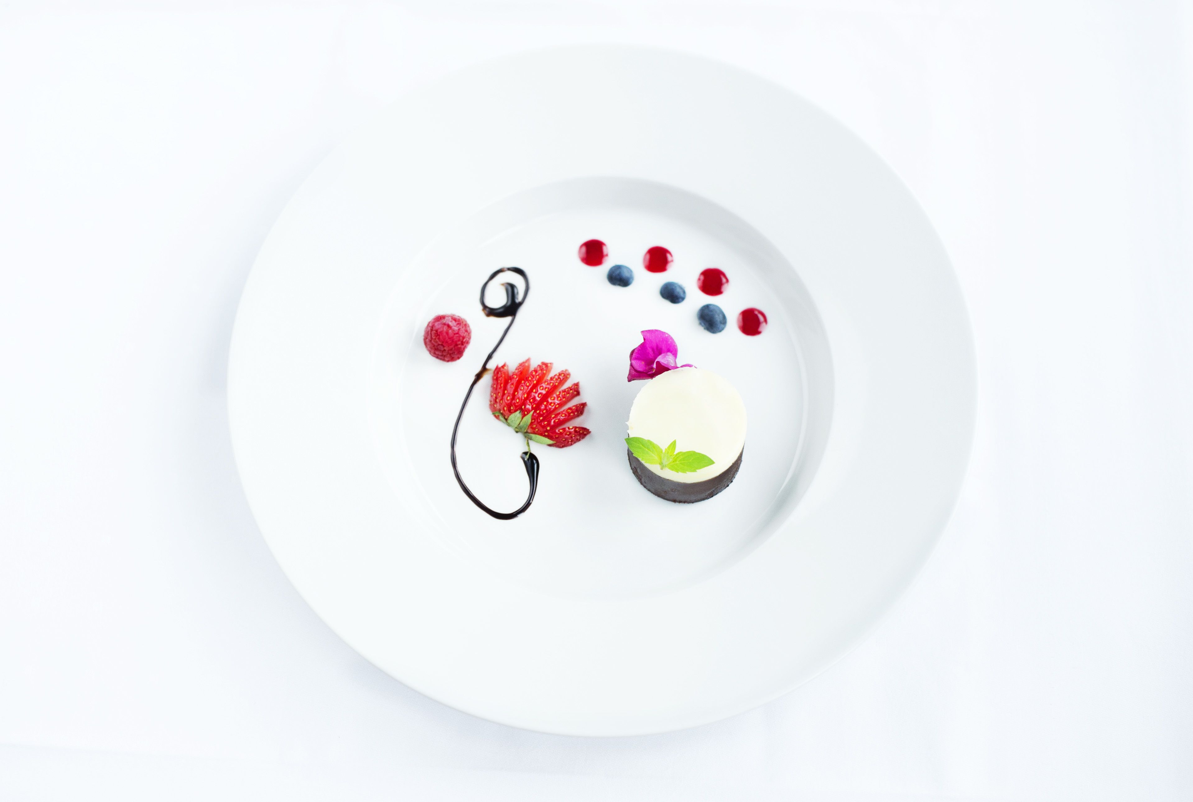Une assiette blanche sur laquelle est présenté un dessert composé d’un nappage au chocolat, de fraises, de myrtilles et d’une feuille de menthe.