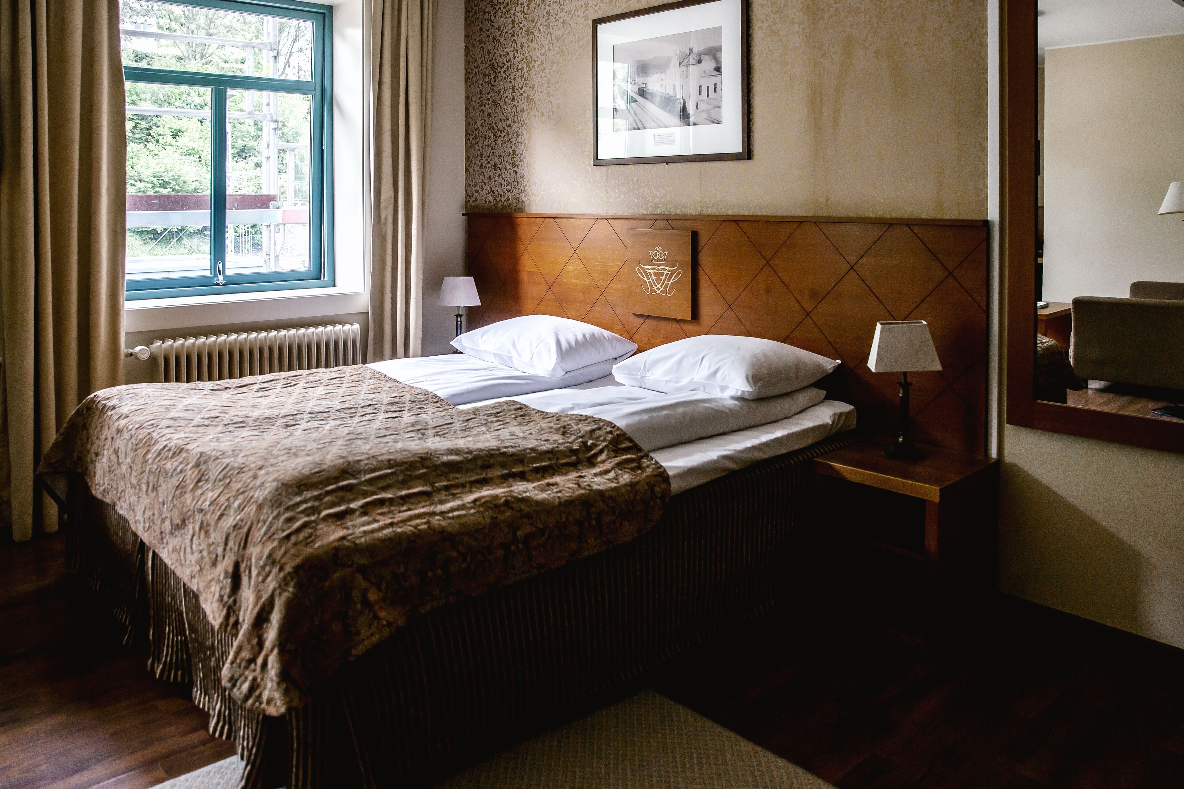 A hotel room with a bed, a nightstand, a lamp, a window with curtains, and a framed picture on the wall.